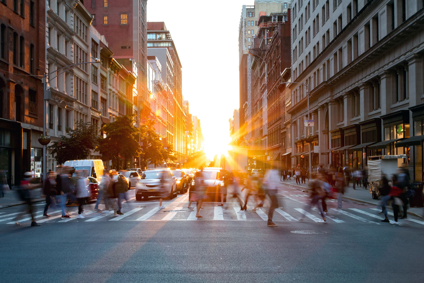 people walking across the street on a crosswalk in a busy city with the sunlight glaring over top of them while cars wait their turn to pass